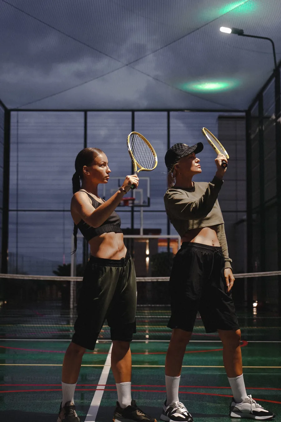 Two players with rackets on an enclosed, floodlit court at dusk